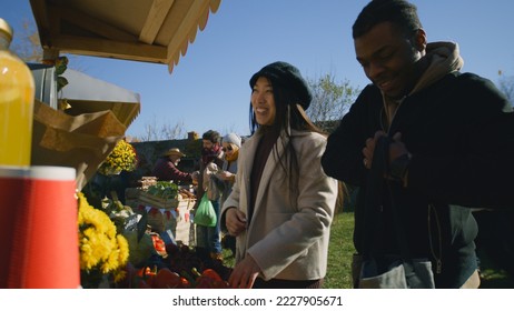 Diverse couple at the stall choosing fruits and vegetables. People walking and shopping on background. Local farmers market or fair outdoors. Vegetarian and organic food. Agriculture. - Powered by Shutterstock