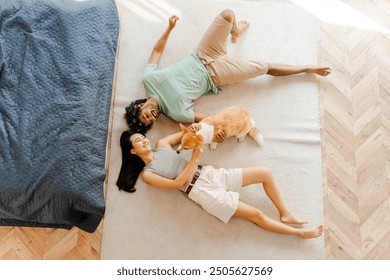 Diverse couple and their playful corgi enjoy quality time at home, lying on a cozy carpet in a stylish bedroom, exuding happiness and love. Relationship concept - Powered by Shutterstock