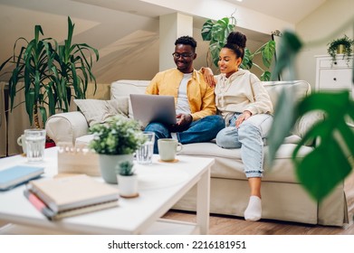 Diverse Couple Using Laptop Together At Home While Sitting Together On A Couch. Smiling African American Man And Hispanic Woman Looking Spending Time Together On Weekend. Modern Interior.