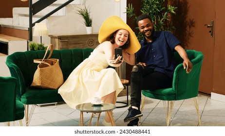 Diverse couple using videocall in hotel lobby, talking to people on online teleconference in lounge area before check in. Tourists enjoying remote video conversation at tropical seaside resort. - Powered by Shutterstock