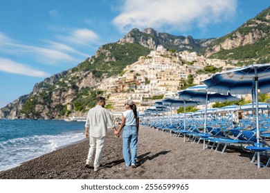 A diverse couple walks hand in hand along a beautiful pebble beach in Positano, Italy, under the warm sun, surrounded by colorful cliffside houses and gentle waves lapping at the shore. - Powered by Shutterstock