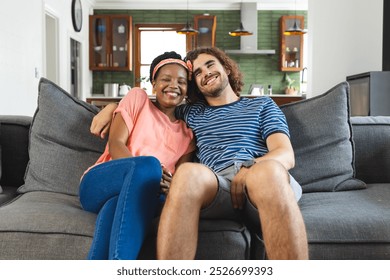 Diverse couple young African American woman and Caucasian man sit closely on a sofa, smiling warmly. Their casual attire and cozy home setting suggest a relaxed and affectionate moment together. - Powered by Shutterstock