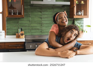 Diverse couple young African American woman embraces a young Caucasian man from behind in a kitchen with copy space. Both are laughing, creating a joyful atmosphere in the bright, modern space. - Powered by Shutterstock