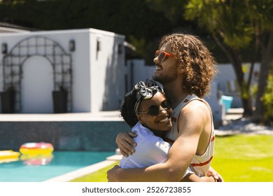 Diverse couple, a young African American woman and Caucasian man, embrace by a pool. Both are smiling, enjoying a sunny day in a backyard setting. - Powered by Shutterstock