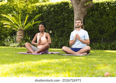 A diverse couple, a young African American woman and a young Caucasian man, are meditating in a serene garden. Both are seated cross-legged on yoga mats, embracing tranquility amidst lush greenery. - Powered by Shutterstock