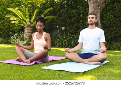 Diverse couple, a young African American woman and Caucasian man, practice yoga outdoors. They are focused on meditation, seated on yoga mats in a serene garden setting. - Powered by Shutterstock