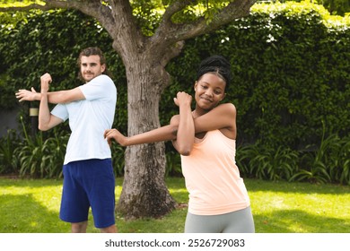 Diverse couple, a young African American woman and Caucasian man, stretch outdoors. They are engaging in a fitness routine in a garden setting. - Powered by Shutterstock