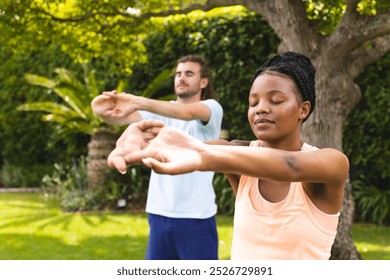 Diverse couple, a young African American woman and Caucasian man, practice yoga outdoors. They are focused on their poses in a serene garden setting, promoting wellness and tranquility. - Powered by Shutterstock