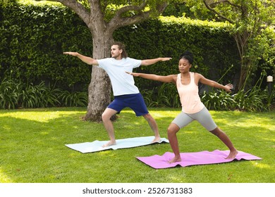 Diverse couple: young African American woman and young Caucasian man practice yoga in a garden. Both are focused on maintaining their poses on yoga mats, surrounded by lush greenery. - Powered by Shutterstock