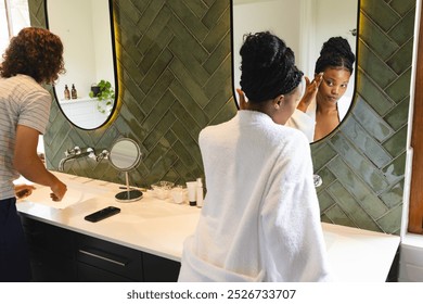 Diverse couple: Young African American woman applies makeup in a bathroom mirror. She wears a white robe, creating a relaxed, personal grooming atmosphere. - Powered by Shutterstock