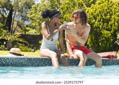 Diverse couple, a young African American woman and young Caucasian man, enjoy drinks by the poolside. She wears a striped swimsuit and headband; he in red shorts, reflecting a relaxed vacation vibe. - Powered by Shutterstock