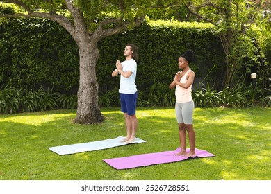 A diverse couple, a young Caucasian man and an African American woman, practice yoga outdoors. Both are standing on yoga mats in a garden, focused on their wellness routine. - Powered by Shutterstock