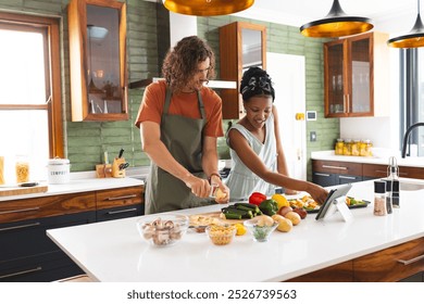 Diverse couple, young Caucasian man and African American girl cooking together in a modern kitchen. He slices vegetables while she follows a recipe on the tablet, both wearing aprons. - Powered by Shutterstock