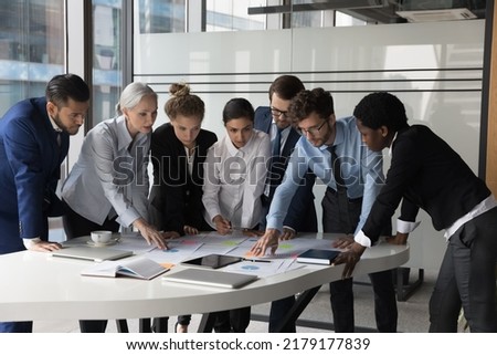 Diverse different aged business group standing at meeting table, negotiating on marketing reports, statistic diagrams, working on startup project together, using scrum approach with sticky notes