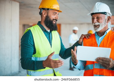 A diverse duo, a construction worker in a yellow helmet and a site manager wearing a white helmet, review plans on a tablet at a construction site, engaged in a cheerful conversation. - Powered by Shutterstock