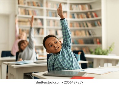 Diverse elementary school students raising their hands to answer teacher questions, sitting at desks in classroom interior. Back to school concept - Powered by Shutterstock