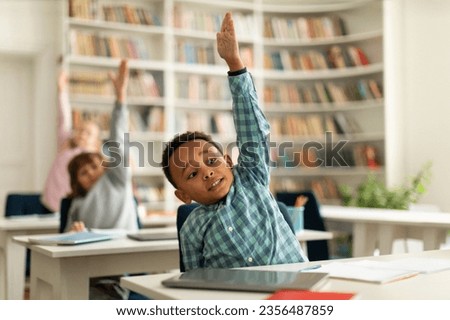 Diverse elementary school students raising their hands to answer teacher questions, sitting at desks in classroom interior. Back to school concept