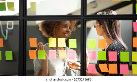 Diverse employees discussing notes on sticky papers, planning project, organizing work, African American and Asian businesswomen talking, sharing ideas, working with stickers on glass wall close up - Powered by Shutterstock