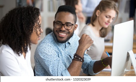 Diverse Employees Working In Shared Office, Focus On Black Smiling Male Talking Flirting With Colleague Sitting At Desk Using Pc. Mentor Provide Information To Trainee Having Friendly Good Relations