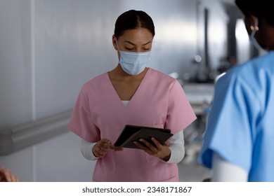 Diverse female doctors wearing face masks using tablet and talking in corridor at hospital. Hospital, communication, teamwork, medicine, healthcare and work, unaltered. - Powered by Shutterstock