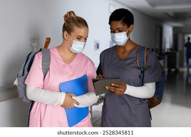 Diverse female doctors wearing face masks discussing work and using tablet in corridor at hospital. Hospital, communication, teamwork, medicine, healthcare and work, unaltered. - Powered by Shutterstock