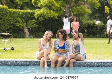 Diverse female friends enjoy a poolside gathering, with a barbecue and friends in the background. The setting suggests a relaxed, social atmosphere during a sunny day. - Powered by Shutterstock