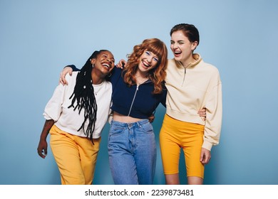 Diverse female friends laughing and having a good time while embracing each other. Group of happy young women enjoying themselves while standing against a blue background. - Powered by Shutterstock