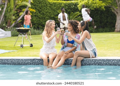 Diverse female friends toast drinks by a poolside, with a diverse group in the background. The setting is a sunny outdoor gathering, a casual social event or a party. - Powered by Shutterstock