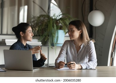Diverse female office employees discussing project, talking at meeting table. Indian mentor explaining work data to intern. Coworkers negotiating on startup, financial report, sharing business ideas