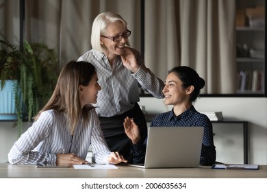 Diverse female office team discussing project and laughing. Business women of different ages and ethnicity talking at workplace with laptop, having fun, sharing good news, enjoying working together