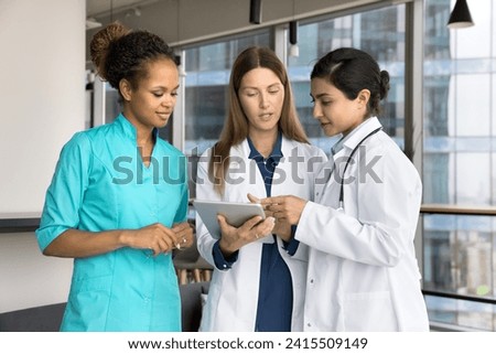 Diverse female team of young doctors discussing medical application on tablet, holding digital gadget, standing and talking in hospital office hall, reviewing patients electronic records
