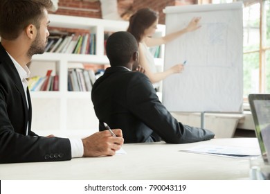 Diverse Focused Businessmen In Suits Listening To Female Coach Giving Presentation On Flipchart Sitting Making Notes At Conference Meeting Table, Corporate Training Business Education Seminar Concept