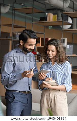 Diverse friendly coworkers talking using digital tablet in office. Indian manager having discussion with latin employee meeting in office lobby discussing project, teaching new employee sharing ideas.