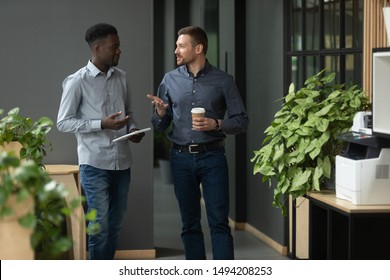 Diverse Friendly Male Partners Colleagues Talking Walking In Modern Office Hallway, Young African American And Caucasian Business Men Discussing Common Project Work Meeting In Company Work Space