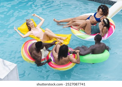 Diverse friends float on colorful rings during pool day. A young Caucasian woman in a pink swimsuit talks with a biracial woman; two African American men and a biracial man relax nearby - Powered by Shutterstock