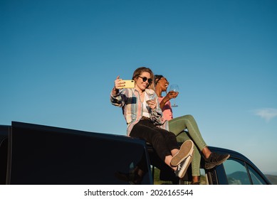 Diverse friends having fun doing video call party celebrating with wine while sitting on top of camper - Focus on left woman face - Powered by Shutterstock