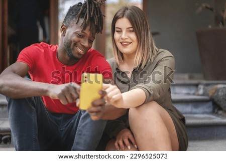 Diverse Friends Sharing Smartphone Content - African man and plus-size woman sitting on outdoor stairs, looking at a smartphone content on a sunny summer day.
