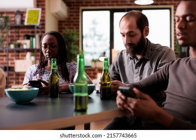 Diverse friends sitting together at table in living room while browsing social media on smartphones. Multiethnic people sitting at home while browsing internet on modern touchscreen devices.