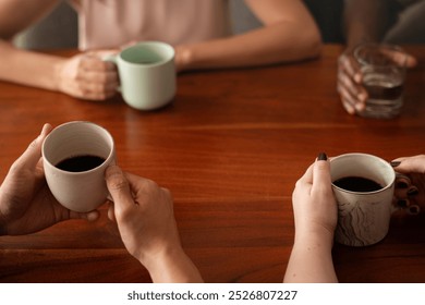 Diverse friends taking a break holding coffee cups and a glass of water. Coffee cups, hands, and wooden table are visible. Coffee cups and hands are the focus. Friends having coffee together at table. - Powered by Shutterstock