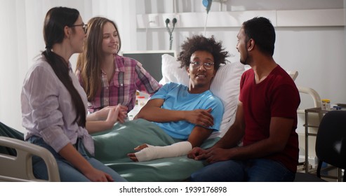Diverse friends visit sick afro patient with broken arm in hospital. Portrait of young afro-american man with hand in plaster cast lying in hospital bed talking with diverse visitors friends