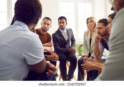 Diverse Group Of Business People Talking During Meeting In Office. Team Of Serious Young Multi Ethnic Men And Women In Suits Sitting In Circle And Listening To Colleague's Suggestion. Teamwork Concept