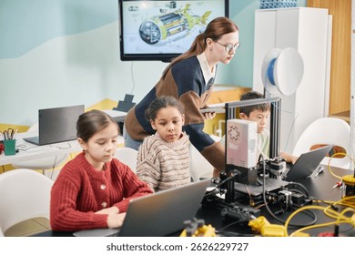 Diverse group of children engaged in coding activities on laptops under guidance of female teacher in tech-enabled classroom with futuristic devices supporting them