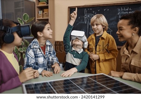 Diverse group of children in modern school classroom with little boy wearing VR headset and pointing up enjoying immersive technology