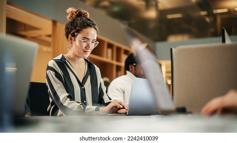 Diverse Group of Creative Colleagues Working on Laptops in Modern Office. Hispanic Female Graphics Designer Smiling and Typing. Colleagues Working on Projects in Background. - Powered by Shutterstock