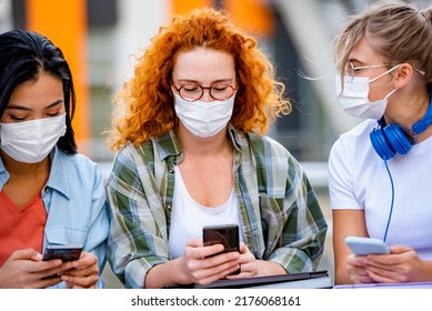 Diverse Group Of Female College Students Sitting In Front Of A University Building Wearing Protective Face Mask, Using Smartphones. 