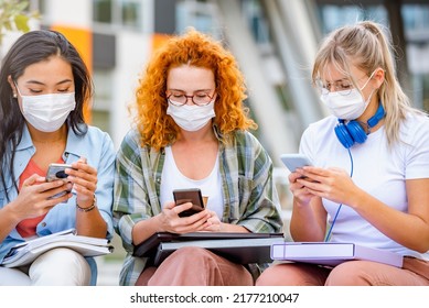 Diverse Group Of Female College Students Sitting In Front Of A University Building Wearing Protective Face Mask, Using Smartphones. 