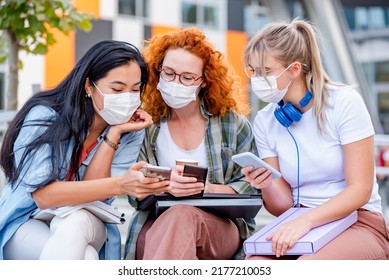 Diverse Group Of Female College Students Sitting In Front Of A University Building Wearing Protective Face Mask, Using Smartphones. 