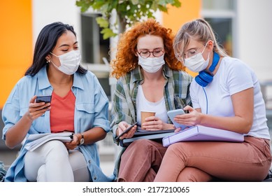 A Diverse Group Of Female College Students Are Sitting In Front Of A University Building Wearing A Protective Face Masks, And Using Smartphones. 