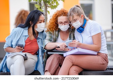 Diverse Group Of Female College Students Sitting In Front Of A University Building Wearing Protective Face Mask, Using Smartphones. 