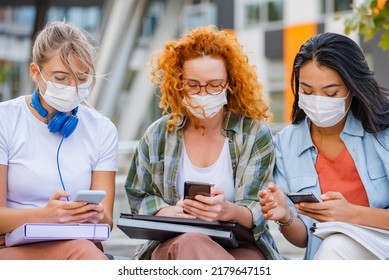 Diverse Group Of Female College Students Sitting In Front Of A University Building Wearing Protective Face Mask, Using Smartphones. 